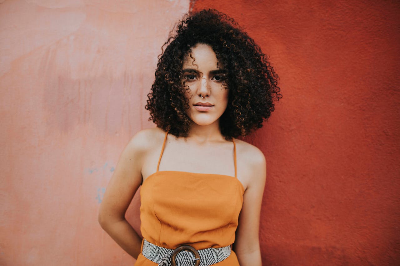Portrait of a fashionable woman with curly hair and nose ring standing against a textured wall.